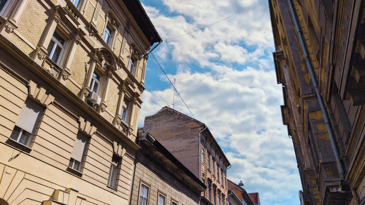 A budapesti hagyományos európai építészet felfelé ívelő látványa, amely elegáns, de időjárásnak kitett történelmi épületeket mutat be részletes homlokzatokkal és díszítőelemekkel. A keskeny utca perspektívája drámai keretet terem